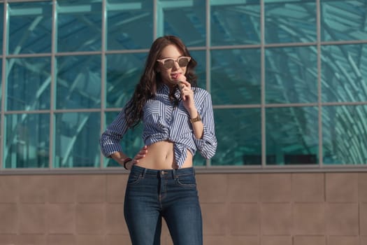 Young, beautiful brunette woman eating a pink ice cream in a waffle horn. She's in a good mood She wears glasses with dark glasses, a shirt in a white and blue strip, and dark blue jeans.