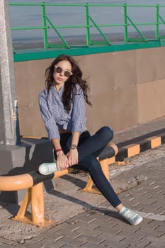 A beautiful girl with sunglasses and torn jeans and a striped shirt. Against the background of large steel structures. Artistic photography. beauty and fashion, style.