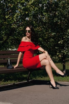 Happy happy young woman in a city park sits on a bench. She is wearing a red dress. Purchase. Beauty, the concept of fashion.