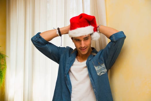 Casual young man celebrating Christmas leaning against a wall smiling happily in his red Santa Claus hat