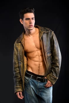 Handsome young man wearing leather jacket on naked torso on dark background, looking away