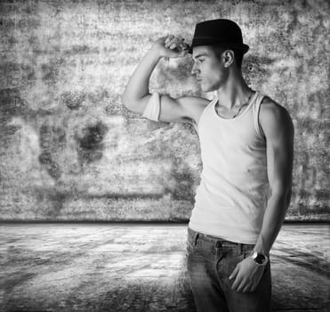 Trendy thoughtful man wearing a fashionable hat and tank top standing in a grunge concrete room staring pensively at the floor