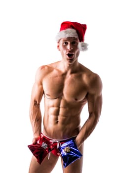 Muscular shirtless young man in a speedo and Santa Claus hat standing holding two colorful festive Christmas gifts to celebrate the season, on white