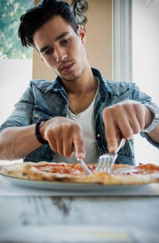 Low angle view of a young man eating an Italian pizza cutting it with a knife and fork
