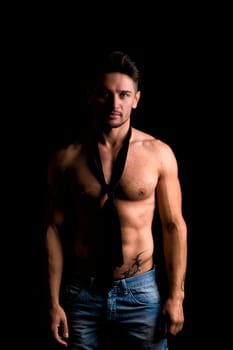 Handsome shirtless muscular man standing on dark background, wearing jeans and neck-tie on naked torso