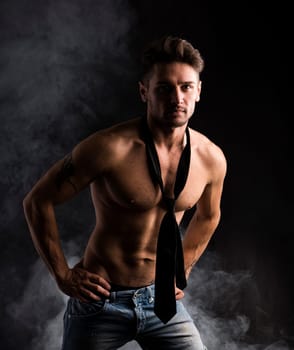 Handsome shirtless muscular man standing on dark smoky background, wearing jeans and neck-tie on naked torso