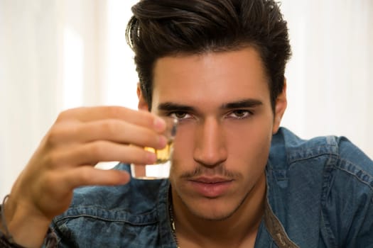 Seductive handsome young man sitting drinking toasting and celebrating with liquor in a shot glass as he gazes at the camera
