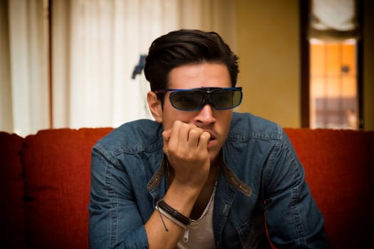 Young man wearing stereoscopic 3d glasses sitting watching a video or television program on a sofa at home sitting forwards with a look of anticipation and his hand to his chin