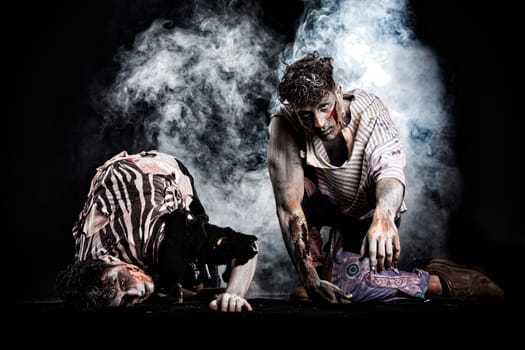 Two male zombies crawling on their knees, on black smoky background, looking at camera. Halloween theme
