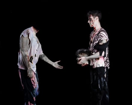 Two male zombies, one giving the head to the decapitated one's body, standing on black background