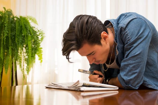 Young man bending over a table at home reading a newspaper with a magnifying glass