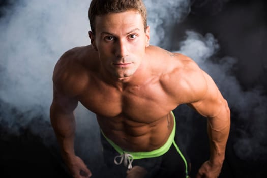 Handsome shirtless bodybuilder shot from above, standing on dark smoky background