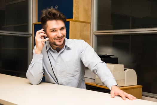 Businessman or customer (support) service man taking a call on his headset phone as he sits at a table in the office looking up to smile at the camera as he listens to the conversation