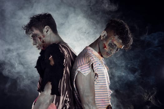 Two male zombies back to back on black smoky background, looking down and away