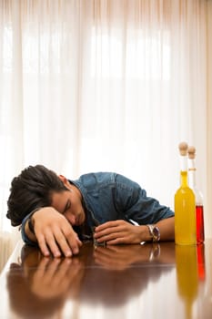 Young man drunk and sleeping alone at a table with two bottles of liquor alongside him sipping from shot glass to drown his sorrows