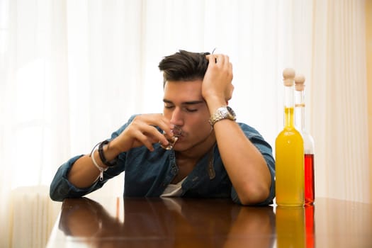 Sleepy, drunk young man sitting drinking alone at a table with two bottles of liquor alongside him sipping from shot glass to drown his sorrows