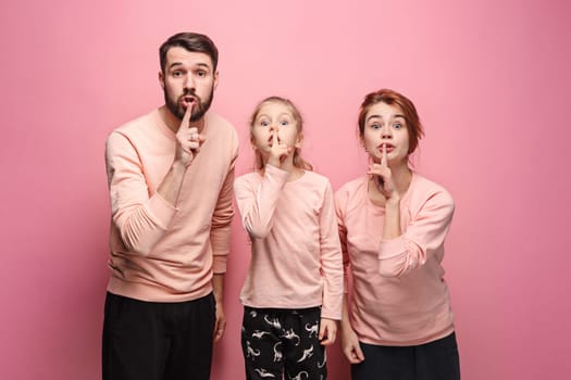 Surprised young family looking at camera on pink studio background and calling for silence by fingers