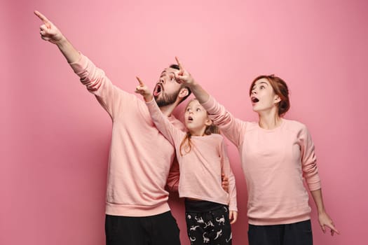 Surprised young family pointing up by fingers on pink studio background