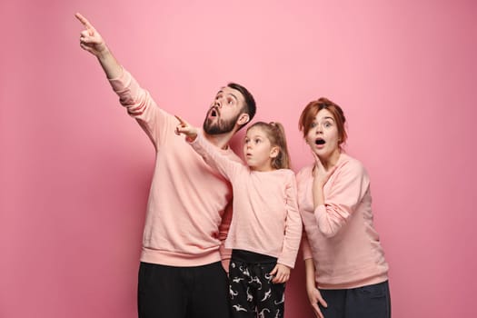 Surprised young family pointing up by fingers on pink studio background