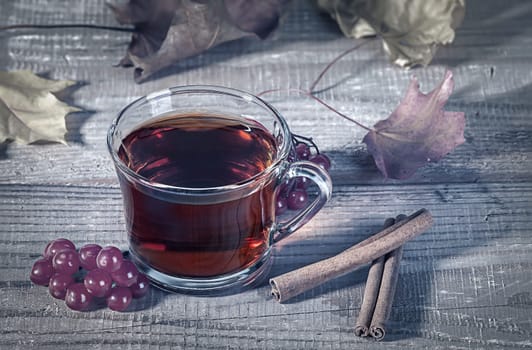 Still life with transparent and fragrant Cup of tea with viburnum and cinnamon on wooden table. Dry autumn maple leaves in the background. Toned photo.