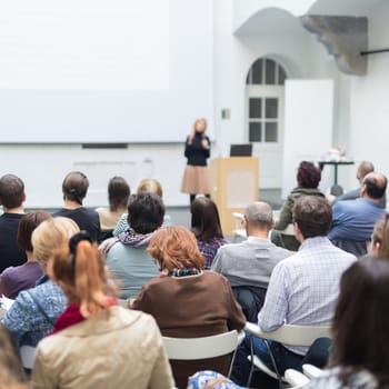 Business and entrepreneurship symposium. Female speaker giving a talk at business meeting. Audience in conference hall. Rear view of unrecognized participant in audience. Copy space on whitescreen.
