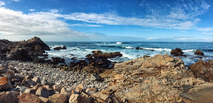 Panorama of Pacific ocean cost in California, USA. Rocky beach in foreground and blue cloudy sky in background.