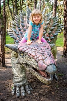 Cute little caucasian girl riding on Ankylosaurus dinosaur in an amusement park in summer
