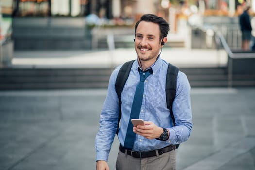 Happy millennial businessman is commuting in Melbourne, Victoria with a smartphone, listening to music through his earphones.