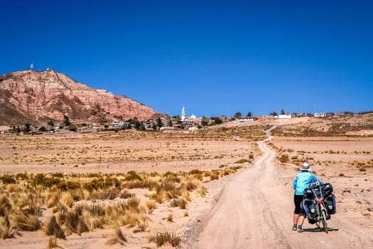 Woman pushing her bicycle on the sandy road in the remote part of Bolivia, approaching small town in the mountains