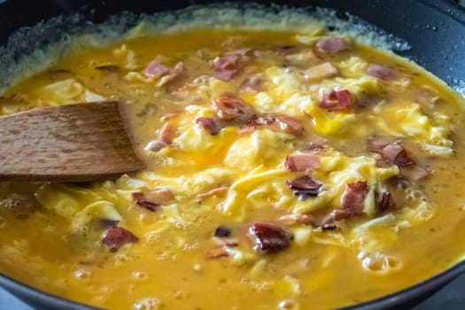 Scrambled eggs with ham and bacon being cooked in a black frying pan, mixed with a wooden ladle