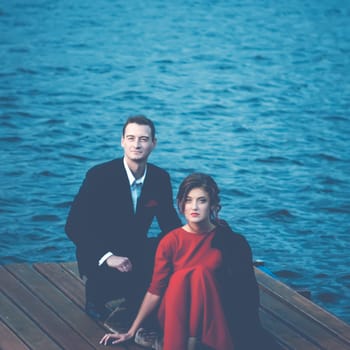 Beautiful couple of lovers - a man in a suit and a woman in a red dress sitting on a pier against a ripple of water. Vintage blue toned filter, square portrait with space for copy.