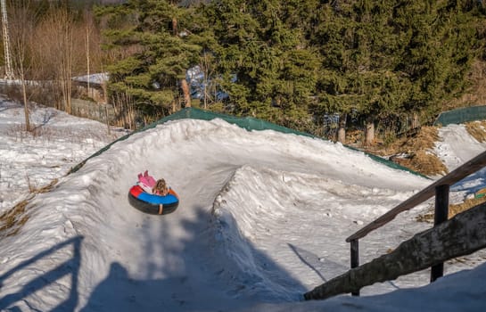 Little girl sledding downhill a snowy and icy hill track on a colorful inflatable tube