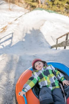 Little boy ready to slide downhill a snowy and icy hill track on a colorful inflatable tube