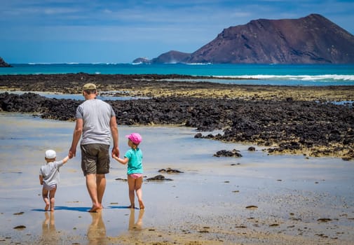 Father with his children, little boy and girl, walking in water waiting for wave to come on the beach in summer