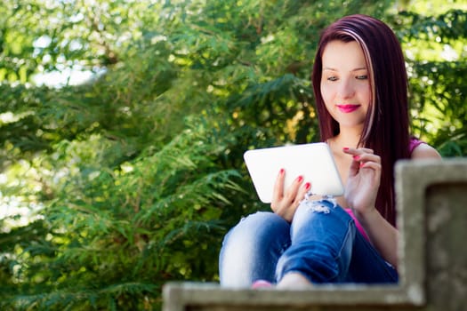 Young woman with tablet is sitting outdoors on stairs in nature.