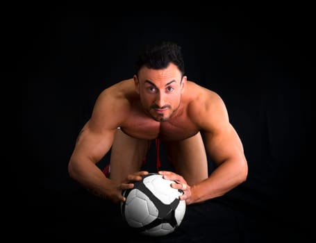 Shirtless muscular man on the floor with soccer ball, isolated on black background