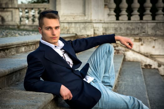 Attractive blond young man sitting on stone stair steps outside, looking at camera
