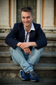 Smiling blond young man sitting on stone stair steps outside, looking at camera