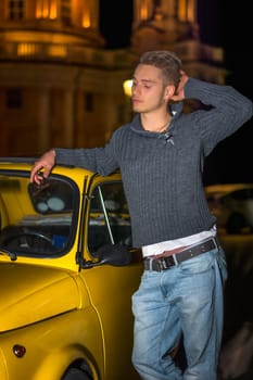 Night shot of handsome blond young man standing next to small yellow car