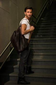 Handsome young man standing with big bag on stairs' steps, looking away