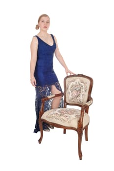 A beautiful young woman standing in a blue long evening gown with.an old antique armchair, isolated for white background.