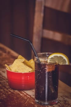Glass containing cola with lemon and small bowl of nachos in the background