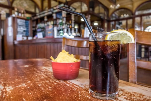 Glass containing cola with lemon and small bowl of nachos in the background