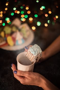 Female hands with cup of tea and christmas cookies house on dark color bokeh lights background