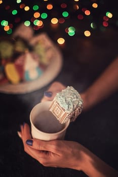 Female hands with cup of tea and christmas cookies house on dark color bokeh lights background