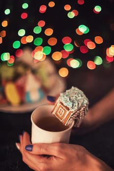 Female hands with cup of tea and christmas cookies house on dark color bokeh lights background