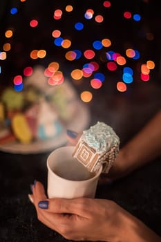 Female hands with cup of tea and christmas cookies house on dark color bokeh lights background