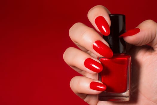 Female hand with red nails is holding red nail polish on red background.