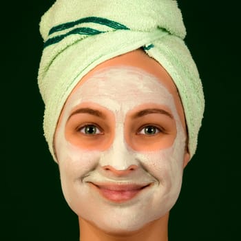 Portrait of woman with face mask and towel on dark green background. Cosmetology concept.