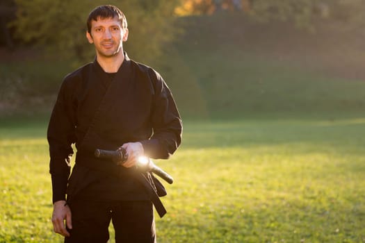 Portrait of smiling ninja in black kimono with sword outdoors in nature.